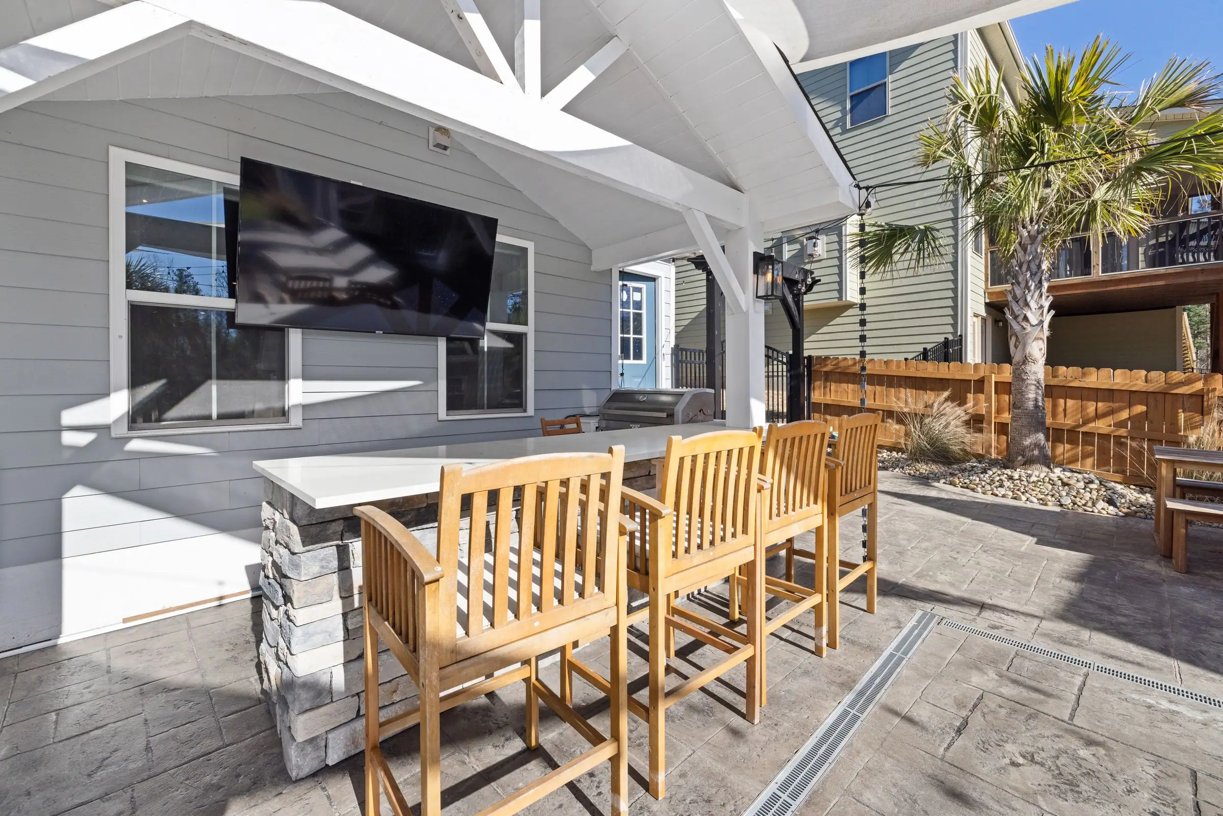 White covered patio with a bar, stools and a wall-mounted TV beside palm trees