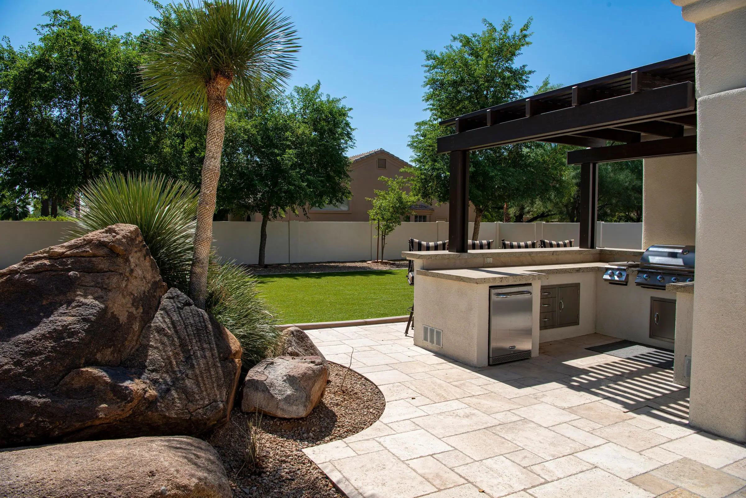 Outdoor kitchen under a dark pergola with desert boulders and a palm tree